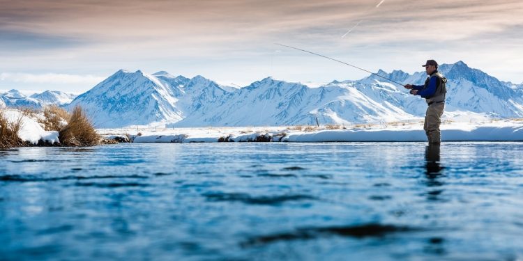A fly fisherman stands in a river with snow-covered mountains standing on the horizon.