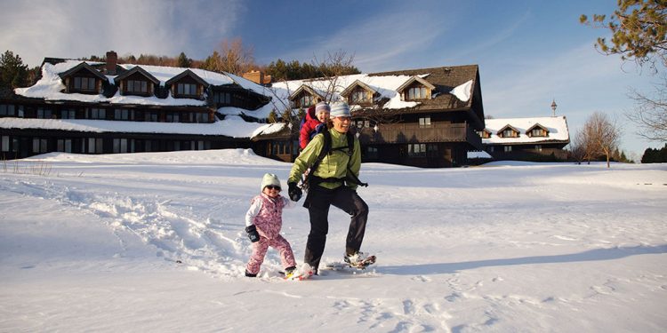 Adult and two kids snowshoeing in front of snow covered chalet.