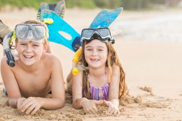 Two siblings - a boy and girl - lie in the sand wearing their snorkeling gear. They have masks and snorkels on their heads and their feet with fins still on behind them kicked up in the air. They are smiling at the camera.