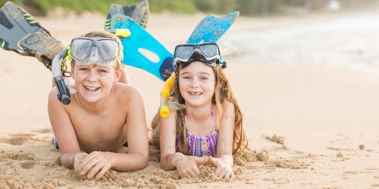 Two siblings - a boy and girl - lie in the sand wearing their snorkeling gear. They have masks and snorkels on their heads and their feet with fins still on behind them kicked up in the air. They are smiling at the camera.