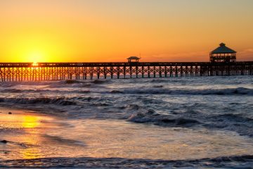 Sun rising behind the Folly pier and reflecting on the beach with a clear sky as waves roll in near Charleston, South Carolina.