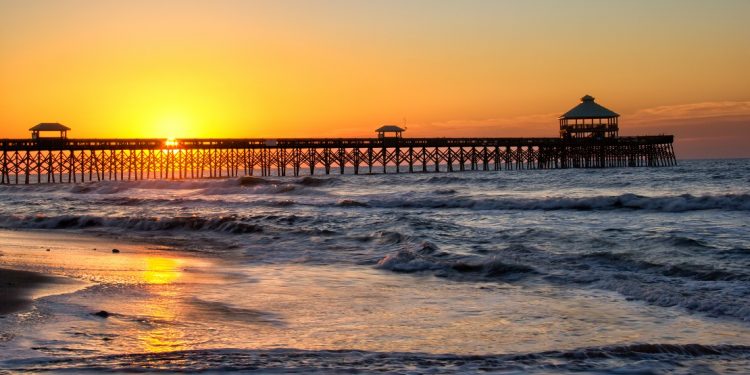 Sun rising behind the Folly pier and reflecting on the beach with a clear sky as waves roll in near Charleston, South Carolina.