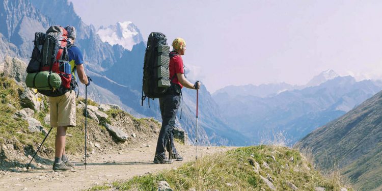 Two backpackers with hiking poles on trail with mountains in background.