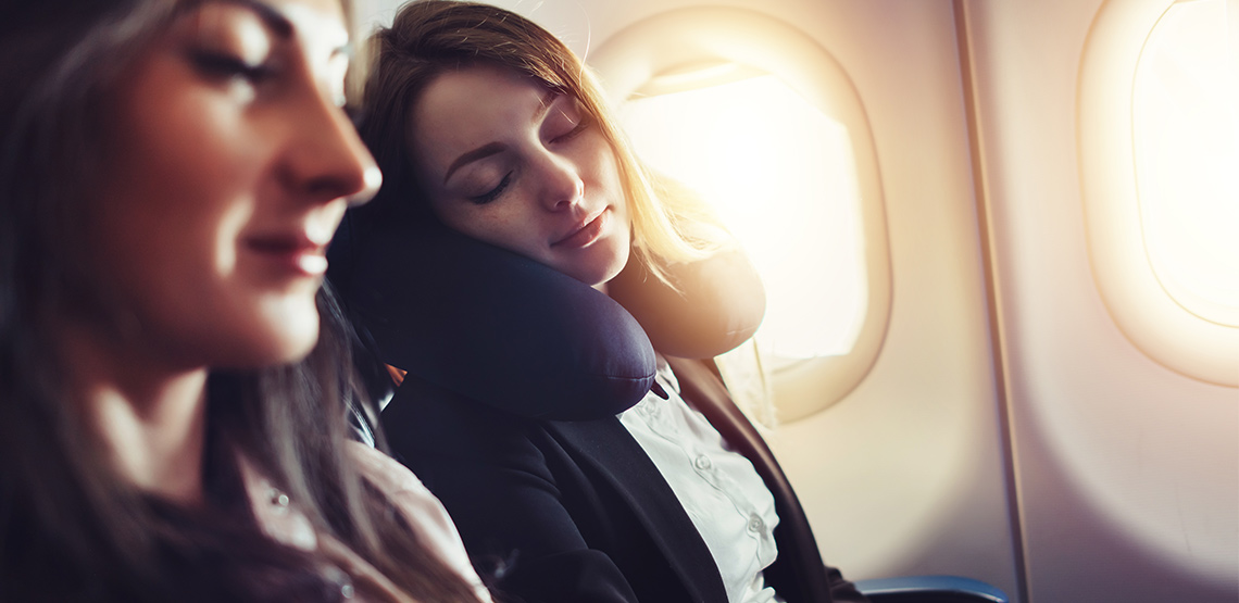 Two women sleeping on a plane, one with a neck pillow.