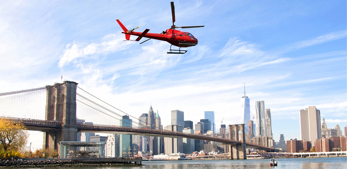 A red helicopter speeds over the Brooklyn Bridge, the skyscrapers of Manhattan visible in the background.