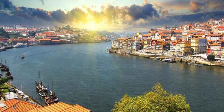 A view of the Douro riverside and Dom Luiz bridge in Porto, Portugal, as the sun sets brilliantly sets off the whitewashed buildings along the shore.