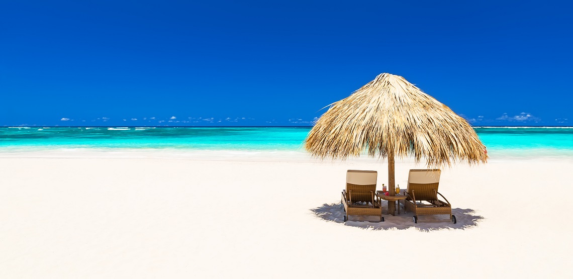 A pair of beach chairs and an umbrella sit on a beautiful, white-sand beach, the deep-blue sky behind them.