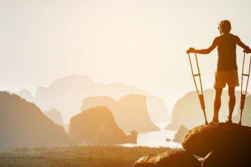 A man holding crutches stands atop of a sun-drenched rock, cliffs rising out of the water in front of him.