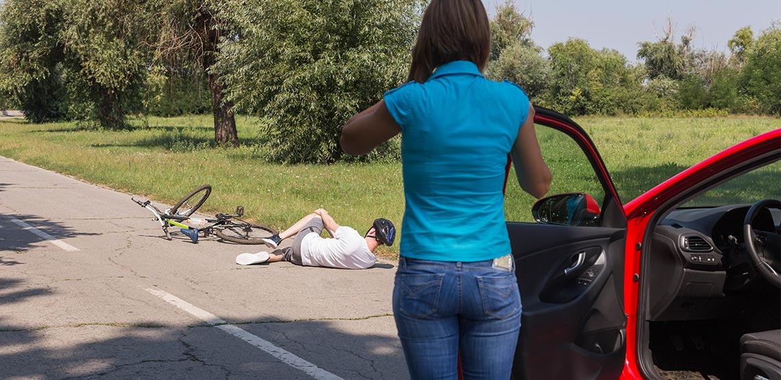 A woman in a blue top stands outside of her car while calling first responders as a cyclist lays on the road clutching his knee.