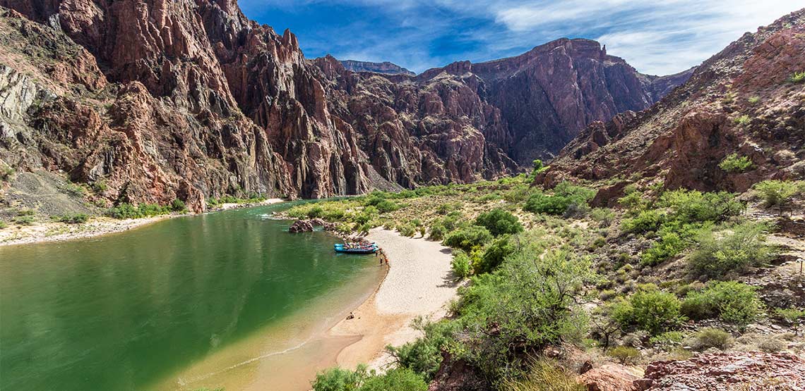 Small rafts rest on a sandy shore by the green Colorado River while the peaks of Grand Canyon cliffs rise around them.