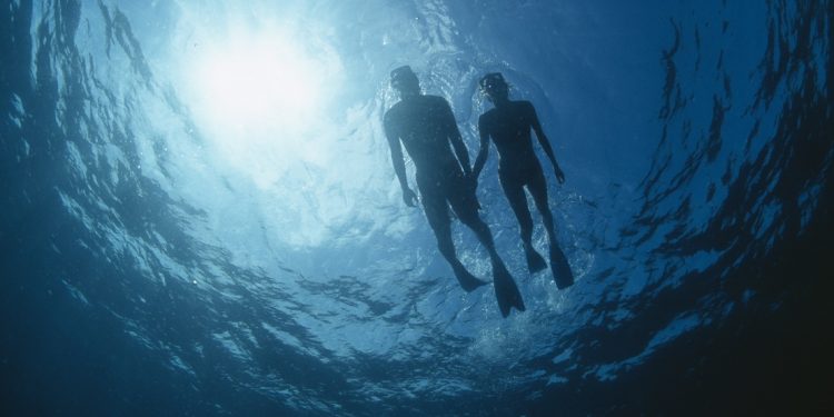 Seen from below, a couple snorkels on top of the waves, the sun a turquoise-blue above them.