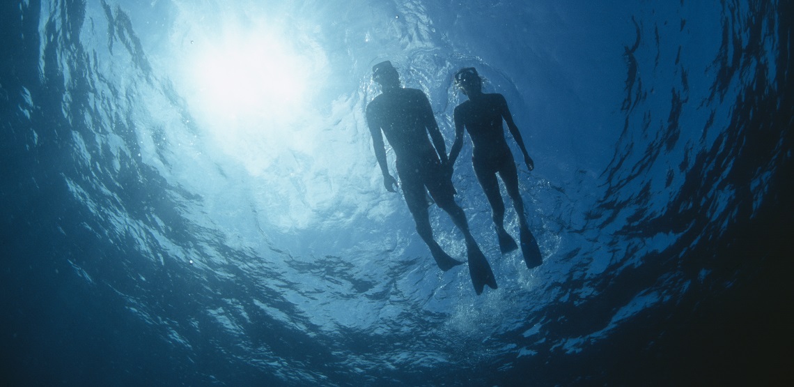 Seen from below, a couple snorkels on top of the waves, the sun a turquoise-blue above them.
