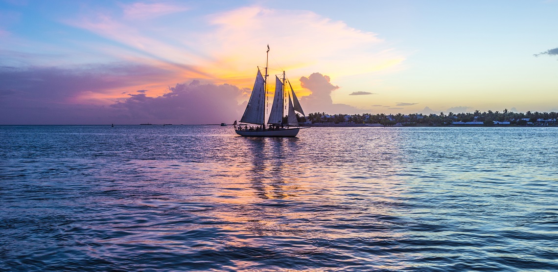 A three-sail sailboat slides by on calm waters at the sunset paints the sky in purples and pinks.