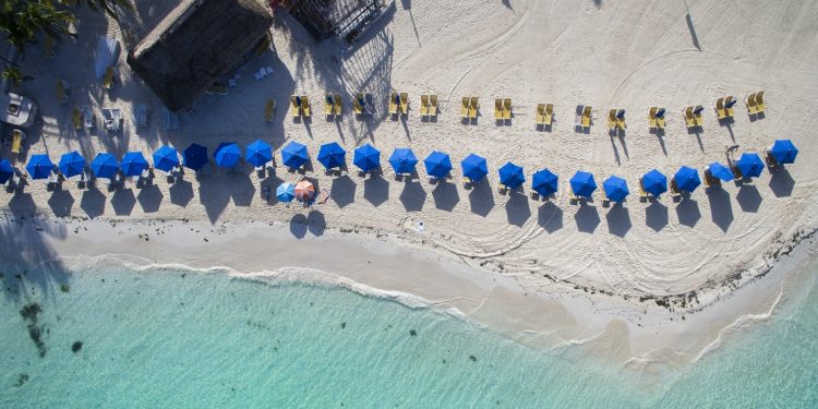 An aerial shot shows blue umbrellas dotting a white-sand beach.