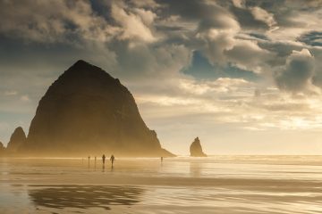 The titanic mass of Haystack Rock towers above the smooth sands of Cannon Beach, Oregon, and beachcombers stroll along the shore.