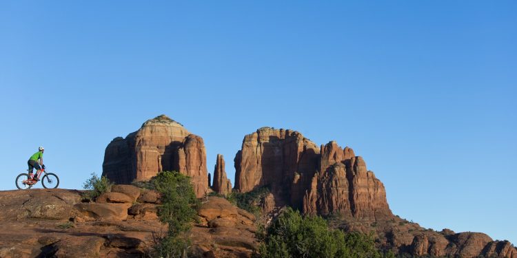 A man rides a popular trail in Sedona, Arizona, USA. He is riding an enduro-style mountain bike. The Cathedral Rock landmark is in the background.
