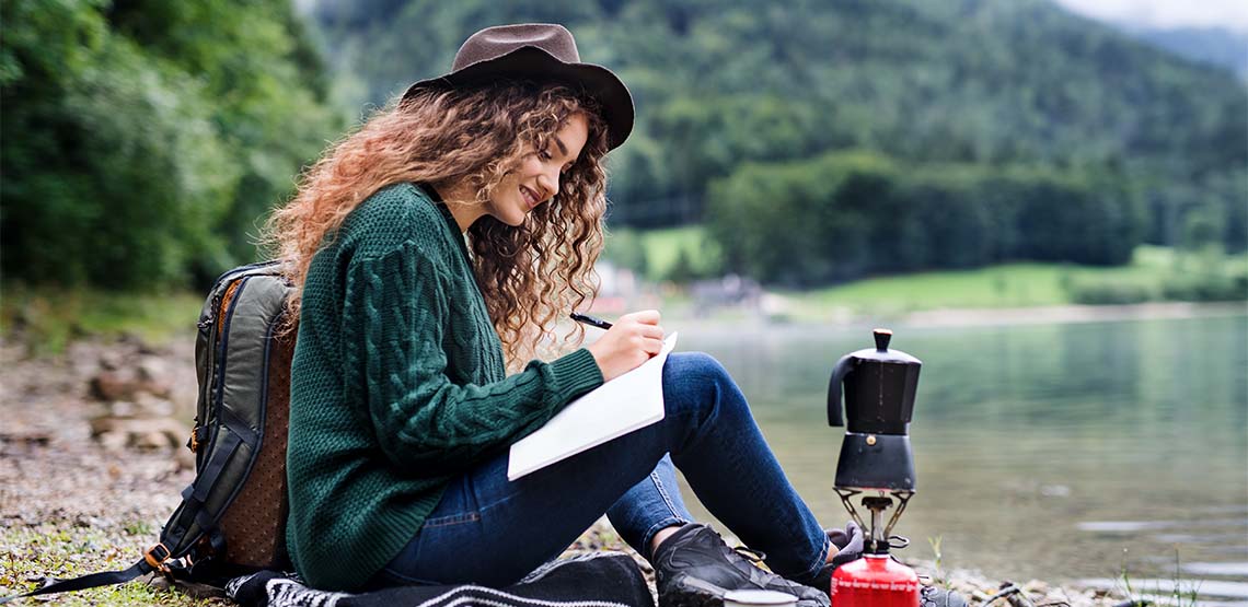 Someone sitting by the water near a forest writing in a travel journal.