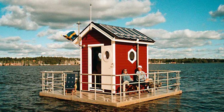 A little read house floating on the water with two people sitting on the deck.