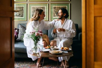 A couple sitting in a hotel room in bathrobes.