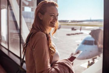 A woman standing near a window holding a plane ticket and smiling.
