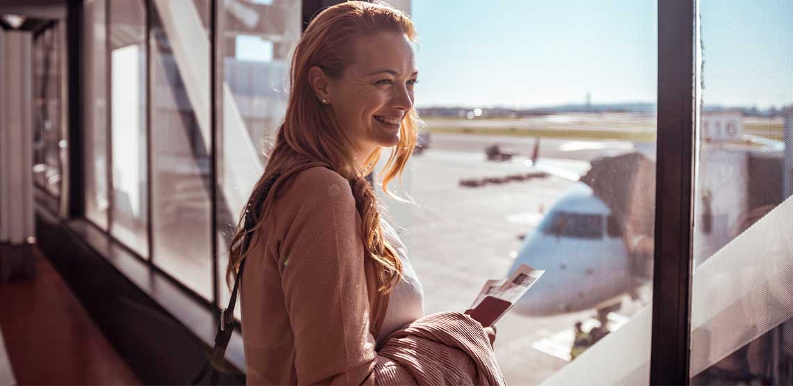 A woman standing near a window holding a plane ticket and smiling.